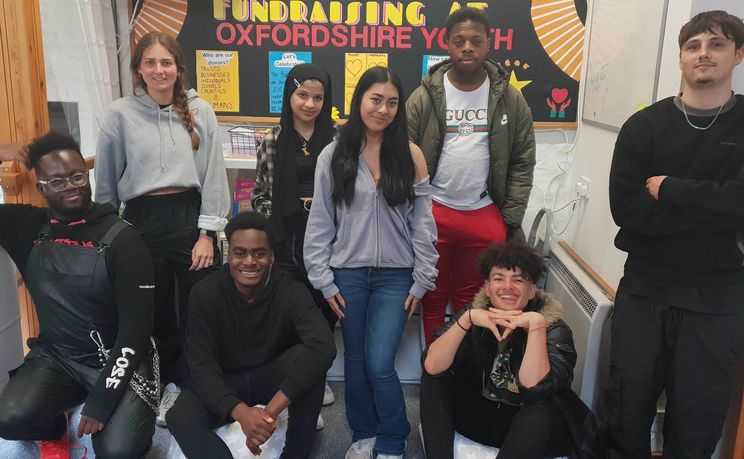 The Youth Awards committee stood together posing and smiling for a photo in front of a colourful fundraising board. From left to right, there is a black male kneeled down, a caucasian female stood up, a black male sat down, a south asian female stood behind him, a south asian female stood in front of her, a black male stood to the right of her, a black and white mixed male sat in the front and a white male stood at the right hand side.